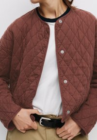 Jachetă matlasată maron închis cu un model în diamant, nasturi cu prindere tip snap argintii și o guler rib elastic negru, purtată cu o cămașă albă și pantaloni kaki.