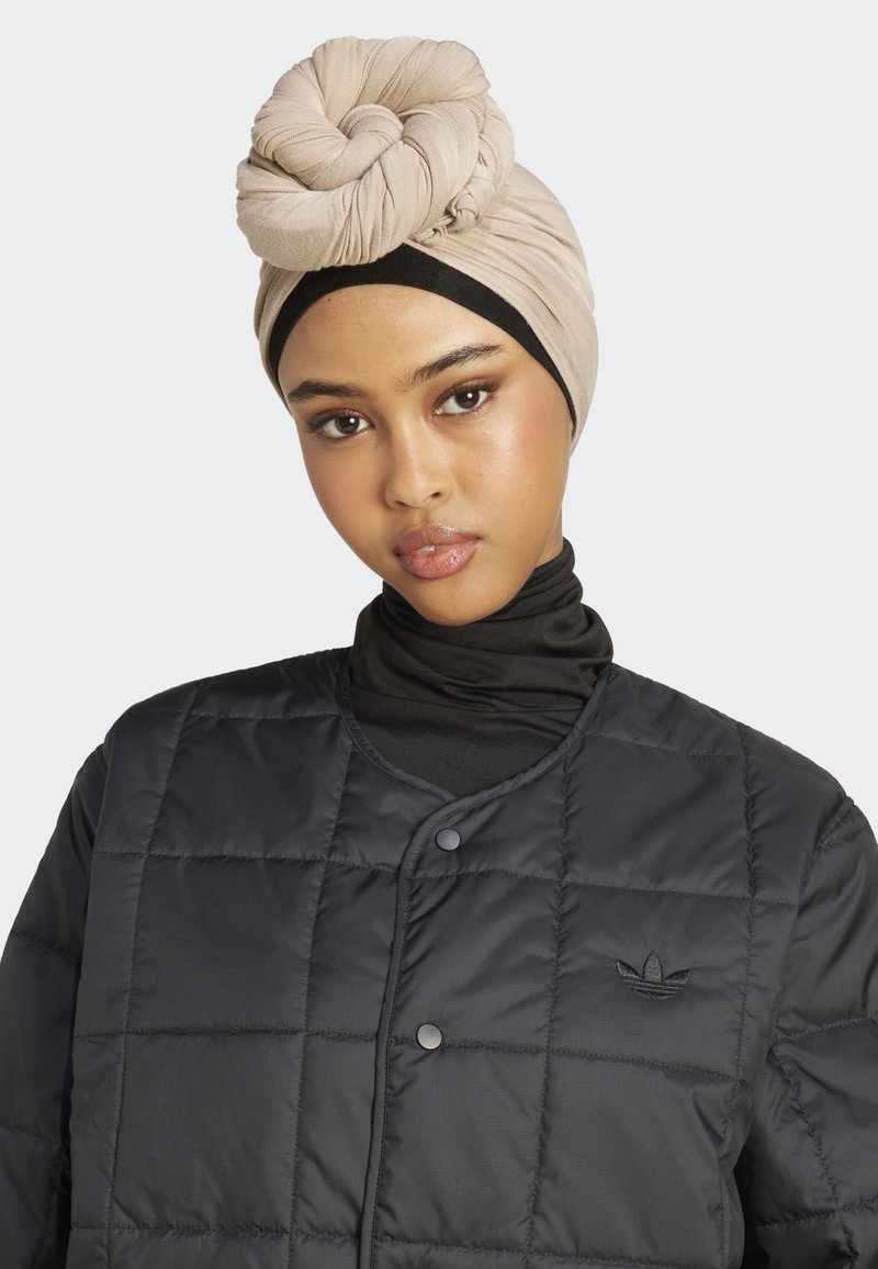 Jeune femme portant un turban beige, un col roulé noir et une veste matelassée noire avec des boutons-pression et un logo sur la poitrine, faisant face à l'avant.