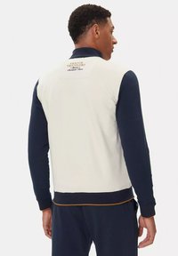 Giacca da uomo con zip, maniche blu navy e corpo crema. Presenta polsini a costine, un logo sulla schiena e un bordo dorato a contrasto in fondo.