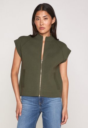 Frau trägt einen olivgrünen, kurzärmligen Zip-Pullover und blaue Jeans, steht vor einem schlichten, hellen Hintergrund.