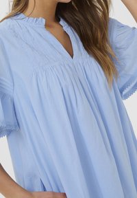 Ljusblå blus i mjukt tyg, med V-ringning, veck och spetsdetaljer på de korta ärmarna. Lätt och flödande design med en avslappnad passform.