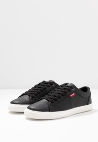 Sneakers in pelle nera con superficie texturizzata, suola in gomma bianca e lacci neri. Presentano un'etichetta con il logo rosso di Levi's sul lato.