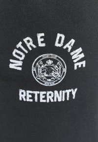 Texte blanc « NOTRE DAME » courbé au-dessus d'un emblème circulaire avec des figures et une couronne, et « RETERNITY » en dessous sur un fond en tissu noir.