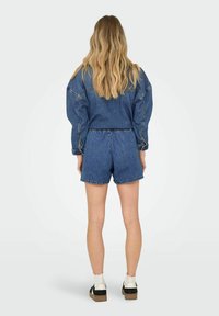 Denim kombinacija sastoji se od široke jakne i odgovarajućih šortsa, oba u srednje plavoj boji. Tkanina je teksturirana s vidljivim šavovima i elastičnom gumicom.