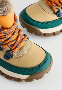 Botte extérieure marron clair et verte avec tige texturée, semelle en caoutchouc, lacets orange et bleus, et col en peluche pour l'isolation.