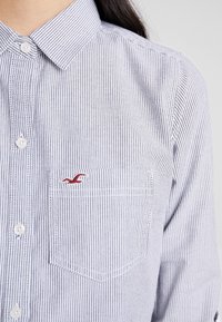 Chemise à rayures bleu et blanc avec une poche avant et un logo rouge. Comprend un col et des boutons blancs.