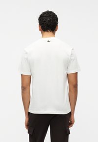 Άνδρας με σγουρά μαλλιά που φοράει απλό λευκό κοντομάνικο μπλουζάκι και μαύρο παντελόνι cargo, γυρισμένος ανάποδα μπροστά σε λευκό φόντο.