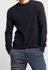 Man som bär en svart texturerad stickad tröja med ribbade ärmar och blågrå byxor, står med händerna i fickorna.