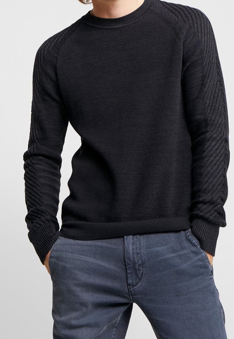 Man som bär en svart texturerad stickad tröja med ribbade ärmar och blågrå byxor, står med händerna i fickorna.