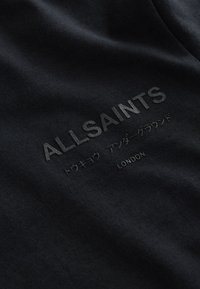 Fekete szövet dombornyomott "ALLSAINTS" felirattal nagy betűkkel, alatta kisebb japán karakterek, és lent "LONDON" nagybetűkkel.