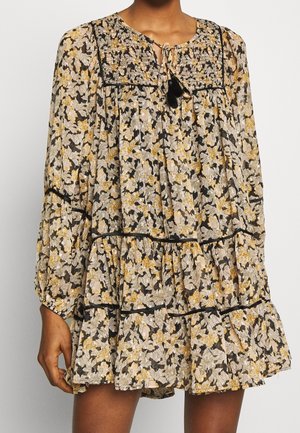 Femme portant une robe ample à motifs floraux avec des manches longues bouffantes, une jupe à volants, un empiècement smocké et un lien à l'avant avec des pampilles dans des tons noir, beige et jaune.