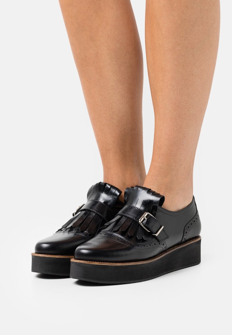 Mocassins à plateforme en cuir noir avec franges et détail de boucle, portés sur jambes nues sur fond blanc.