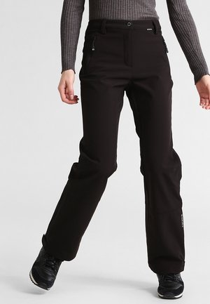 Persona con pantalones de exterior negros y zapatillas negras, de pie con las piernas ligeramente separadas frente a un fondo liso.