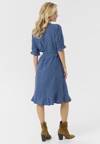 Modra cvetna obleka s kratkimi rokavi, volanastim robom in pasom na vezanje. Ujemajo se z rjavimi gležnjači. Gladko teksturo tkanine.