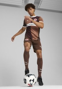 Maillot de football marron et noir avec des accents de logo, manches courtes, et shorts assortis. Le joueur se tient avec un ballon de soccer noir et blanc.