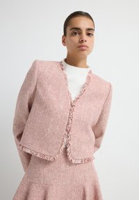 Chaqueta de tweed roja y blanca con bordes deshilachados, hombros estructurados y un diseño corto. Llevada sobre una camiseta blanca.