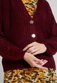 Burgundy knitted cardigan with a v-neck, featuring two silver buttons and textured sleeves. Worn over a patterned yellow and brown dress.