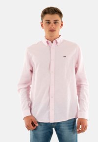 Camisa de botones de manga larga color rosa claro con un cuello tradicional, que presenta botones blancos y un pequeño logo en el lado izquierdo del pecho.