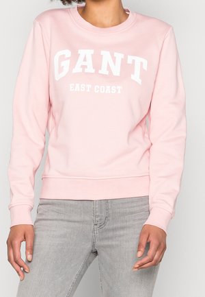 Person trägt ein hellrosa GANT-Sweatshirt mit dem Schriftzug "EAST COAST" und hellgraue Jeans, steht vor einem einfarbigen Hintergrund.
