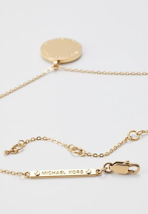 Collier chaîne en or avec une plaque rectangulaire gravée de "MICHAEL KORS" et un pendentif rond sur fond blanc.