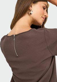 Donna con lunghi capelli castani indossa orecchini a cerchio in argento e un top marrone a texture con cerniera sul retro, che guarda di lato su uno sfondo semplice.