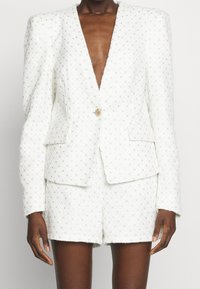 Blazer en tweed blanc avec un col en V profond, orné d'accents dorés et d'une finition texturée. Associé à un short taille haute assorti.
