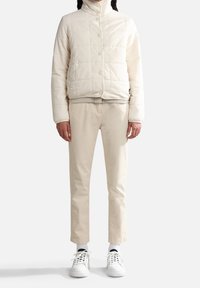 Cremefarvet kviltet pufferjakke med høj krave, parret med lysebeige bukser og hvide sneakers. Glat tekstur og knaplukninger.