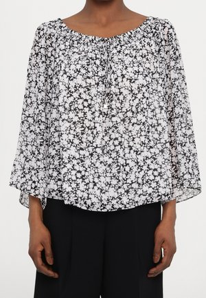 Blouse fluide fleurie noir et blanc avec manches trois-quarts et col rond, portée avec un pantalon noir.