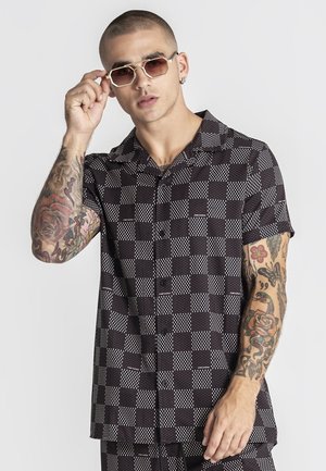 Muž s tetovaniami si nastavuje geometrické slnečné okuliare, oblečený v čiernobielej krátkoročnej košeli s gombíkmi a zladených nohaviciach.