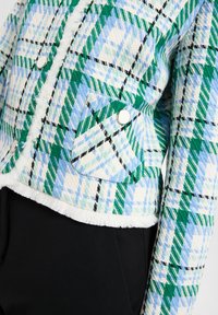 Geblokte jas in blauwe, groene en witte tinten met zwarte accenten. Getextureerde stof, franjes aan de onderkant en ronde knopen op de zakken.