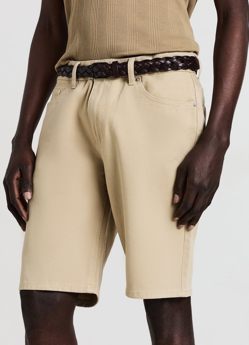 Shorts in cotone beige con taglio dritto e texture sottile, dotati di tasche laterali e una cintura in pelle intrecciata scura in vita.