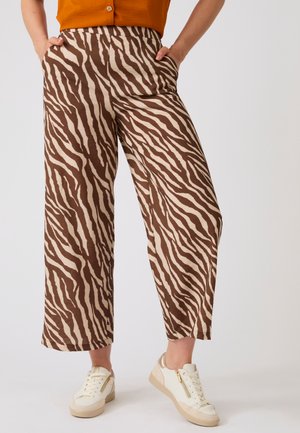 Persona che indossa pantaloni larghi a righe zebrate marroni e beige, sneakers bianche con suole beige e una maglia arancione con le mani nelle tasche.