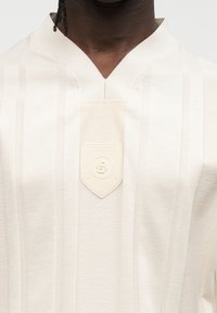 Πουκάμισο σε κρεμ χρώμα με κάθετες ρίγες, γιακά σε σχήμα V και κεντρικό υφασμάτινο τμήμα με ασπίδα και λογότυπο.