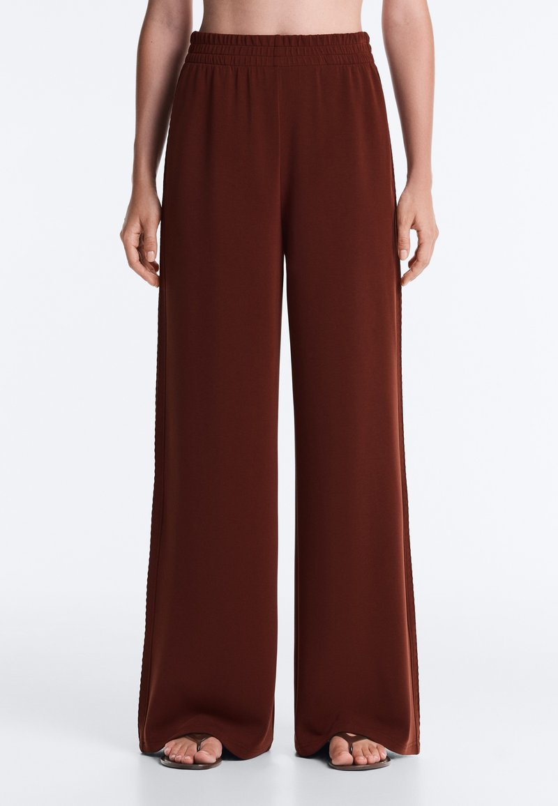 Femme portant un pantalon marron taille haute à jambes larges avec une ceinture élastique, associée à des sandales noires tongs, debout devant un fond uni.