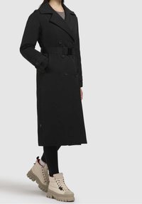 Femme portant un long trench-coat noir ceinturé, des leggings noirs et des bottes beige épaisses à lacets sur un fond uni.