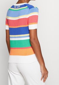 Kvinna som bär en kortärmad stickad topp med horisontella flerfärgade ränder i blått, korall, grönt, orange och vitt, matchat med vita byxor.