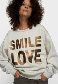 Mujer con cabello rizado vistiendo una sudadera gris claro con un gran texto metálico en bronce que dice "SMILE LOVE", con las manos en los bolsillos.