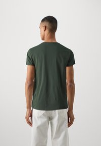 Grünes Baumwoll-T-Shirt mit kurzen Ärmeln und Rundhalsausschnitt, von hinten betrachtet, getragen mit weißen Jeans. Glatte Textur, lässige Passform.