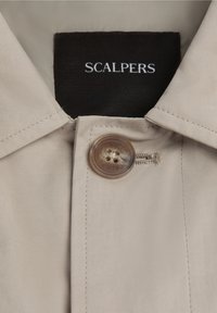 Materialul paltonului bej are un finisaj satinat. Prezintă un nasture brown și eticheta "SCALPERS" în alb pe un fundal întunecat.