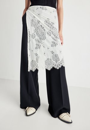 Persona vestida con pantalones negros de pierna ancha, falda envolvente blanca de encaje floral y zapatos de punta blancos con tachuelas, de pie contra un fondo liso.