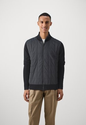 Padded zip-up jacket features a quilted gray front and black knitted sleeves. Front pocket visible with ribbed cuffs and hem.