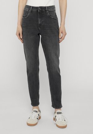 Sorte højtkwaistede smalle jeans iført hvide Adidas sneakers, set forfra fra talje til fødder på en ensfarvet baggrund.