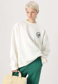 Krēmkrāsas sporta krekls mīkstā audumā, ar apļveida logo uzlīmi. Apvienots ar zaļām strukturētām biksēm un haki krāsas somu ar divām rokturiem.