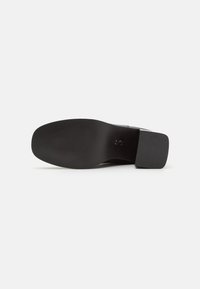 Suela de zapato de tacón negro con una superficie plana y texturizada y un leve tacón, con un sutil logo grabado cerca del área del arco.