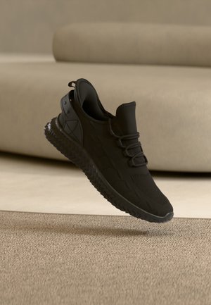 Baskets athlétiques noires avec semelle texturée et lacets, flottant au-dessus d'un tapis beige devant un canapé beige clair.