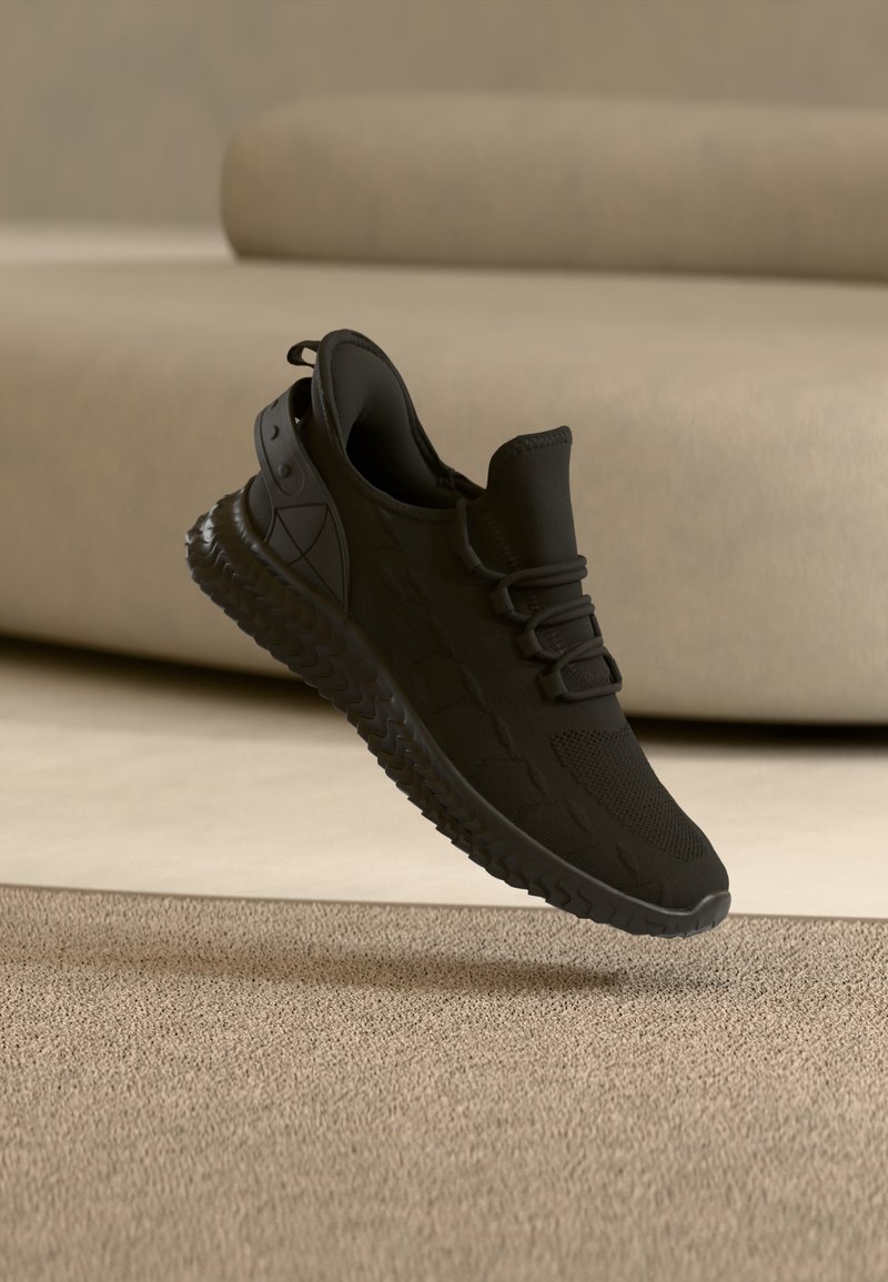 Μαύρο αθλητικό παπούτσι με υφή στη σόλα και κορδόνια, που αιωρείται πάνω από μπεζ χαλί μπροστά από ανοιχτό μπεζ καναπέ.