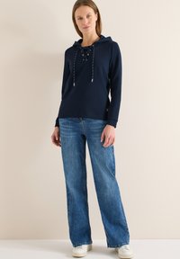 Pull-over bleu marine avec un devant à lacets et une capuche, assorti à un jean en denim bleu clair ample et des baskets blanches. Texture lisse.