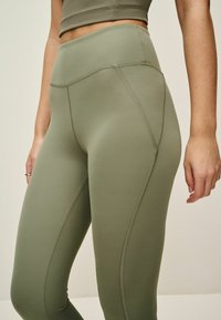Magas derekú zöld leggings sima, rugalmas anyagból. Oldalt varrás részletek találhatók, amelyek javítják a illeszkedést és a modern dizájnt biztosítanak.