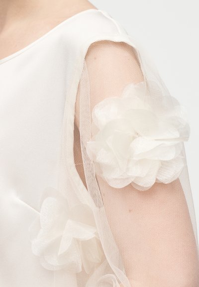 Διάφανο άσπρο τούλινο μανίκι με μεγάλες υφασμάτινες εφαρμογές λουλουδιών, που φοριέται πάνω σε γυμνό δέρμα σε λευκό σατέν φόρεμα.