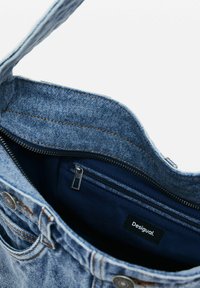 Denim-Umhängetasche mit blauer Außenseite, ausgestattet mit einer reißverschlossenen Innentasche und einem schwarzen Etikett. Sichtbare Nähte und weiche Textur.